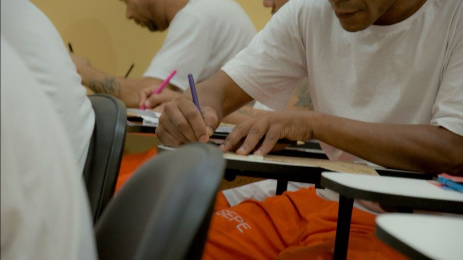 A foto mostra um ambiente de sala de aula onde vários homens, de pele morena e calvos ou com cabelo raspado, estão sentados em carteiras escolares, escrevendo ou estudando. Em primeiro plano, um homem com uma camiseta branca e calça laranja de uniforme (com a inscrição "SUSEPE" na perna) escreve concentrado em um caderno com uma caneta roxa, suas mãos e braços tatuados. Ao fundo, outros homens, também de camiseta branca, realizam a mesma atividade, alguns com canetas coloridas. O ambiente é simples, com paredes claras, e a cena foca na atividade de aprendizado e concentração dos indivíduos.