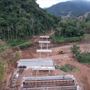 I will describe each image individually for accessibility purposes.

Image 1: Executado com recursos do Fundo Estadual de Apoio ao Desenvolvimento dos Pequenos Estabelecimentos Rurais.jpg

Esta é uma fotografia em perspectiva de um grande auditório lotado, onde um homem está discursando no primeiro plano.

No primeiro plano, à esquerda e de costas para o observador, um homem branco com barba, vestindo uma camisa branca de manga comprida e calça jeans escura, segura um microfone preto com a mão direita e gesticula com a esquerda. Ele parece estar falando para a plateia. À sua frente, em uma mesa de madeira, há alguns papéis e uma pilha de livros ou pastas verdes.

Atrás do orador e ocupando a maior parte da imagem, está uma vasta plateia composta por centenas de pessoas sentadas em cadeiras. A maioria das pessoas na plateia é jovem, e algumas estão com celulares erguidos, possivelmente gravando ou tirando fotos. Ao fundo da plateia, há uma cortina azul escura e, acima dela, um mural branco com algumas frases e logotipos. Uma das frases visíveis no mural diz "SOLIDARIEDADE AO POVO GAÚCHO".

O teto do auditório é branco e alto, com diversas luzes e um projetor visível. A iluminação geral é clara. No canto inferior esquerdo, parte de outro homem com cabelos grisalhos e óculos, vestindo um blazer escuro, é visível sentado, de perfil.

Image 2: Ação PFPS Cigarros.jpg

Esta é uma imagem em close-up que mostra a parte traseira de um caminhão de carga, com sua carroceria aberta revelando uma vasta quantidade de caixas de papelão empilhadas do chão ao teto. As caixas estão alinhadas em várias prateleiras dentro do caminhão, ocupando todo o espaço visível.

A maioria das caixas é marrom clara e parece estar selada com plástico transparente. Algumas das caixas têm a palavra "BOX" impressa em letras maiúsculas escuras, com as palavras "CUIDADO FRÁGIL" acima e abaixo dela. Em outras caixas, há um logotipo azul e branco com a bandeira do Brasil e a frase "INDÚSTRIA BRASILEIRA".

No canto inferior esquerdo da imagem, uma pequena parte da cabine do caminhão é visível, mostrando os pneus dianteiros e o logotipo "ICH". Uma escada de madeira, parecendo ser usada para acessar as prateleiras superiores do caminhão, está encostada na lateral da carroceria. O chão embaixo do caminhão é de asfalto escuro. A iluminação na imagem é fraca, indicando que a foto foi tirada em um ambiente interno ou à noite.

Image 3: Governo deve entregar nova ponte de Sinimbu até o fim de 2025.jpg

Esta é uma fotografia aérea de um canteiro de obras de uma ponte em um vale verdejante, com montanhas e um rio ao fundo.

No primeiro plano e plano médio, estruturas de concreto cinza que formam os pilares e as lajes de uma ponte estão visíveis em várias etapas de construção. As bases dos pilares estão fincadas no leito de um rio estreito de água escura, que serpenteia pelo vale à direita da ponte. Materiais de construção, como madeira e escoramentos, estão espalhados pelo solo de terra batida marrom-avermelhada ao redor das estruturas. Uma escavadeira amarela está visível à direita das fundações da ponte.

O vale é densamente arborizado, com encostas verdes cobertas por árvores altas e folhagem exuberante. Algumas árvores têm troncos mais claros e finos. Ao fundo, as montanhas são cobertas por vegetação e há uma névoa que as envolve. O céu está nublado, com tons de branco e cinza.
