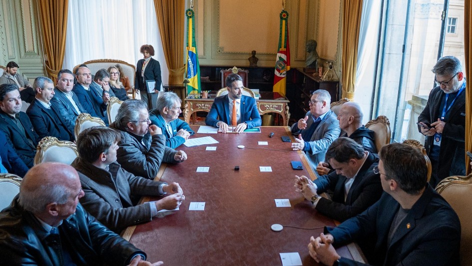 Sala imponente com teto alto e janelas grandes; bandeiras do Brasil e outra entidade ao fundo. Pessoas sentadas ao redor de uma longa mesa retangular discutem documentos e ideias, lideradas por alguém na cabeceira com postura de autoridade. Ambiente transmite seriedade e contexto governamental.