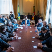 Sala imponente com teto alto e janelas grandes; bandeiras do Brasil e outra entidade ao fundo. Pessoas sentadas ao redor de uma longa mesa retangular discutem documentos e ideias, lideradas por alguém na cabeceira com postura de autoridade. Ambiente transmite seriedade e contexto governamental.