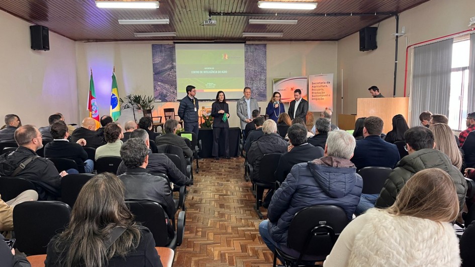 Fotografia colorida de um auditório durante um evento do Centro de Inteligência do Agronegócio. O local está cheio, com diversas pessoas sentadas assistindo a uma apresentação. Ao fundo, seis pessoas estão de pé em frente a um telão que exibe o título “Centro de Inteligência do Agro”. Ao lado do telão, há bandeiras do Brasil e do Rio Grande do Sul, além de banners institucionais. O ambiente tem piso de parquet e forro de madeira. A maioria dos presentes veste roupas de frio, como casacos e jaquetas.