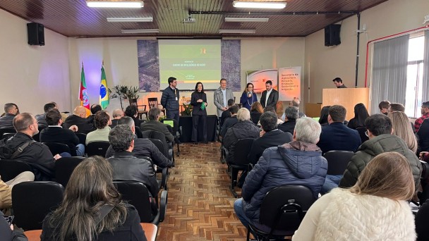 Fotografia colorida de um auditório durante um evento do Centro de Inteligência do Agronegócio. O local está cheio, com diversas pessoas sentadas assistindo a uma apresentação. Ao fundo, seis pessoas estão de pé em frente a um telão que exibe o título “Centro de Inteligência do Agro”. Ao lado do telão, há bandeiras do Brasil e do Rio Grande do Sul, além de banners institucionais. O ambiente tem piso de parquet e forro de madeira. A maioria dos presentes veste roupas de frio, como casacos e jaquetas.