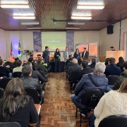 Fotografia colorida de um auditório durante um evento do Centro de Inteligência do Agronegócio. O local está cheio, com diversas pessoas sentadas assistindo a uma apresentação. Ao fundo, seis pessoas estão de pé em frente a um telão que exibe o título “Centro de Inteligência do Agro”. Ao lado do telão, há bandeiras do Brasil e do Rio Grande do Sul, além de banners institucionais. O ambiente tem piso de parquet e forro de madeira. A maioria dos presentes veste roupas de frio, como casacos e jaquetas.