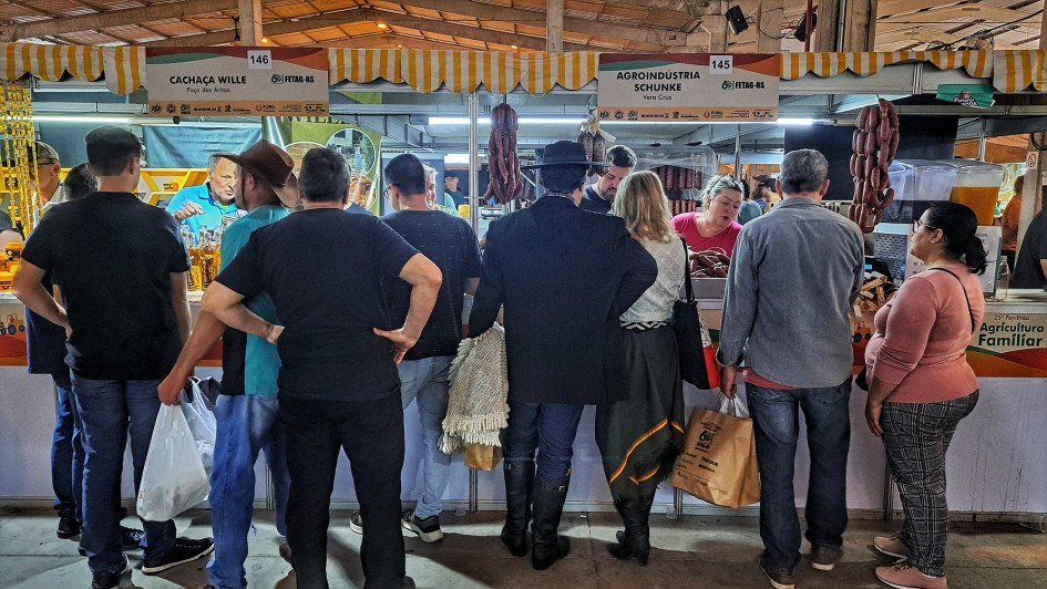 A foto mostra uma feira movimentada, com diversas pessoas de costas, observando os produtos em estandes iluminados. Os estandes têm toldos listrados em amarelo e branco. Ao fundo, pendem vários tipos de embutidos e carnes, indicando que são barracas de produtos alimentícios. As pessoas, em sua maioria adultos, estão vestidas com roupas casuais e interagem com os vendedores. A iluminação clara destaca os produtos expostos. O chão é de concreto, e a cena transmite a atmosfera vibrante de um mercado de alimentos artesanais. A feira está repleta de visitantes de todas as idades, sugerindo um ambiente de comércio e lazer.