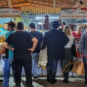 A foto mostra uma feira movimentada, com diversas pessoas de costas, observando os produtos em estandes iluminados. Os estandes têm toldos listrados em amarelo e branco. Ao fundo, pendem vários tipos de embutidos e carnes, indicando que são barracas de produtos alimentícios. As pessoas, em sua maioria adultos, estão vestidas com roupas casuais e interagem com os vendedores. A iluminação clara destaca os produtos expostos. O chão é de concreto, e a cena transmite a atmosfera vibrante de um mercado de alimentos artesanais. A feira está repleta de visitantes de todas as idades, sugerindo um ambiente de comércio e lazer.