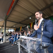 A imagem mostra um evento oficial com a presença do governador do Rio Grande do Sul, Eduardo Leite, que está em destaque à direita, falando ao microfone em um púlpito transparente. Ele veste terno azul e gravata laranja. Ao fundo, há uma plateia composta por autoridades civis e militares, algumas fardadas, sentadas em um palanque coberto. A decoração inclui tecidos nas cores da bandeira do Rio Grande do Sul (verde, vermelho e amarelo). Também é possível ver jornalistas, fotógrafos e cinegrafistas registrando o momento. O evento acontece em um espaço aberto, com público ao fundo, sugerindo uma cerimônia pública, possivelmente de formatura ou entrega de viaturas à Brigada Militar.