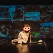 Em um palco escuro, uma pessoa está sentada no chão, com o corpo parcialmente coberto por um pano branco e usando um grande capacete ou máscara que representa um rosto infantil com olhos grandes e expressão neutra. A máscara tem cabelo cacheado castanho. Ao fundo, há uma grande projeção com várias imagens de animais, aparentemente cachorros em diferentes ambientes. A iluminação destaca a figura no centro do palco, criando um contraste dramático com o fundo. A cena remete a uma apresentação teatral ou performance artística.