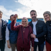 A imagem mostra um grupo de seis pessoas em frente a uma casa de cor azul, em um momento de celebração. No centro da cena, uma senhora idosa, vestindo um conjunto esportivo bordô e um lenço na cabeça, sorri e ergue um molho de chaves com uma miniatura de casa, simbolizando a conquista de uma moradia. Ao seu lado direito está o governador do Rio Grande do Sul, Eduardo Leite, sorridente, com a mão apoiada no ombro dela. À esquerda, estão dois homens — um deles usando colete azul com brasão do Estado. À direita, uma mulher loira de casaco preto também sorri. Todos demonstram alegria e empatia, em um clima de gratidão e conquista. A entrega ocorre ao ar livre, com céu parcialmente nublado.