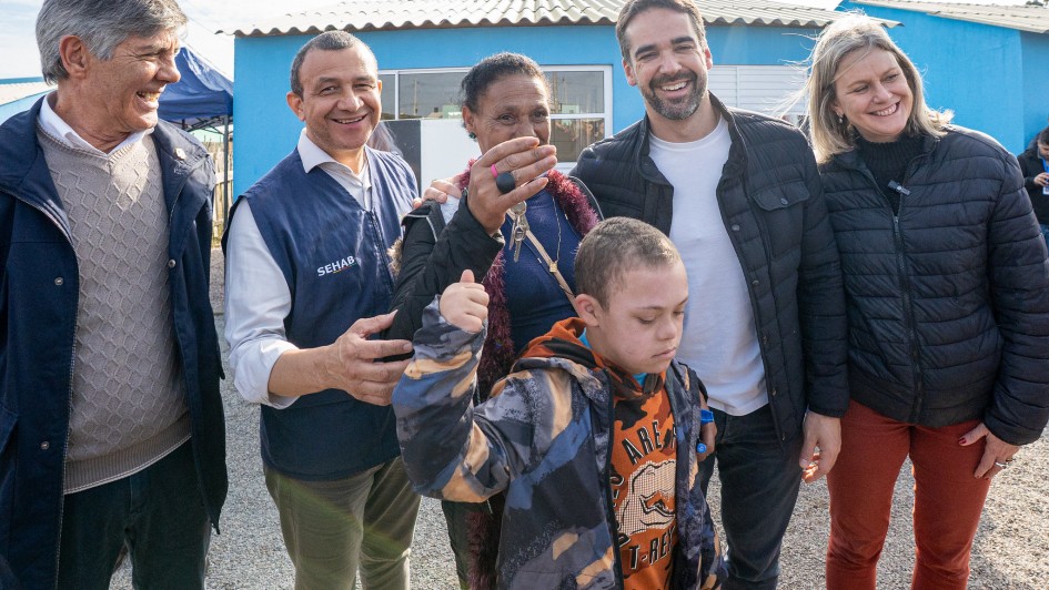 Seis pessoas estão reunidas sorrindo ao lado de uma mulher que segura as chaves da nova casa. Em primeiro plano, um menino com síndrome de Down veste um moletom camuflado e faz um gesto com o braço. Ao fundo, casas azuis e uma tenda azul são visíveis. Todos celebram a entrega da moradia.