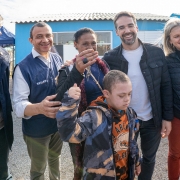 Seis pessoas estão reunidas sorrindo ao lado de uma mulher que segura as chaves da nova casa. Em primeiro plano, um menino com síndrome de Down veste um moletom camuflado e faz um gesto com o braço. Ao fundo, casas azuis e uma tenda azul são visíveis. Todos celebram a entrega da moradia.