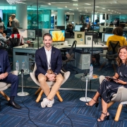 Imagem em um ambiente interno com mesas e computadores. O governador Eduardo Leite está posicionado em destaque, ao lado de um homem e uma mulher. Os três estão sentados em poltronas e sorriem para a câmera. 