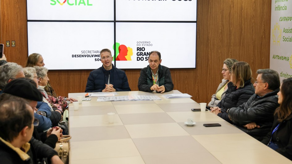 Em uma sala de reuniões, cerca de 14 pessoas participam de um encontro ao redor de uma mesa retangular. Dois homens, sentados ao centro, lideram a reunião. Atrás deles, há painéis com as logomarcas "RS Social", "Reconstruir OSCs", "Secretaria de Desenvolvimento Social" e "Governo do Estado do Rio Grande do Sul". À direita, vê-se parte da parede com palavras como "primeira infância", "direitos" e "autonomia".