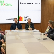 Em uma sala de reuniões, cerca de 14 pessoas participam de um encontro ao redor de uma mesa retangular. Dois homens, sentados ao centro, lideram a reunião. Atrás deles, há painéis com as logomarcas "RS Social", "Reconstruir OSCs", "Secretaria de Desenvolvimento Social" e "Governo do Estado do Rio Grande do Sul". À direita, vê-se parte da parede com palavras como "primeira infância", "direitos" e "autonomia".