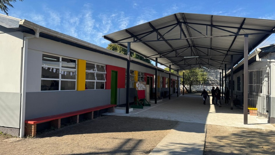 A foto mostra um corredor externo da EEEF Doutor Ottoni Xavier, com prédios baixos de um andar à esquerda e à direita. As fachadas dos prédios são pintadas em tons de cinza, amarelo, vermelho e verde, adicionando cor ao ambiente escolar. Uma cobertura metálica com estrutura aparente protege o corredor central, que é de concreto. Um banco vermelho longo está junto à parede da esquerda. Ao fundo, algumas pessoas circulam. O céu é azul com algumas nuvens, e árvores fornecem sombra, criando um ambiente funcional e colorido.