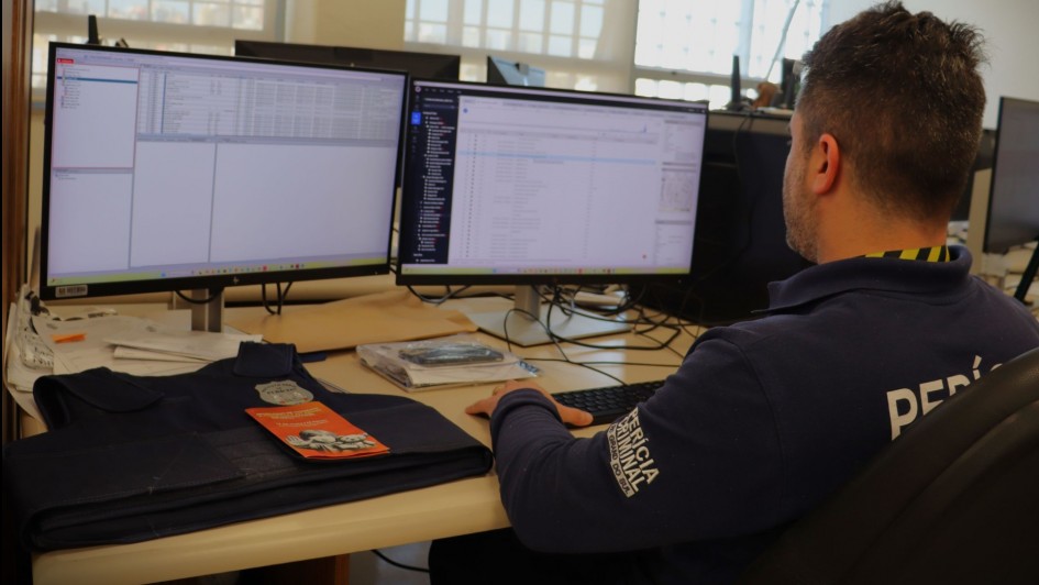 A foto mostra um homem, visto de costas, sentado em uma cadeira de escritório e concentrado em dois monitores de computador que exibem linhas de código e dados. Ele veste uma camiseta polo azul marinho com as palavras "PERÍCIA CRIMINAL" parcialmente visíveis nas costas. Sobre a mesa, há uma pasta, papéis e outros objetos de escritório. A luz natural entra por janelas gradeadas ao fundo. A cena sugere um ambiente de trabalho de investigação ou análise de dados, típico de perícia.