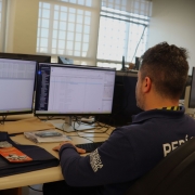 A foto mostra um homem, visto de costas, sentado em uma cadeira de escritório e concentrado em dois monitores de computador que exibem linhas de código e dados. Ele veste uma camiseta polo azul marinho com as palavras "PERÍCIA CRIMINAL" parcialmente visíveis nas costas. Sobre a mesa, há uma pasta, papéis e outros objetos de escritório. A luz natural entra por janelas gradeadas ao fundo. A cena sugere um ambiente de trabalho de investigação ou análise de dados, típico de perícia.