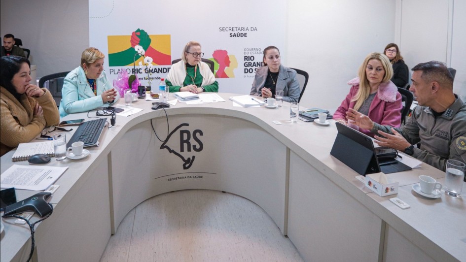 A foto mostra um grupo de oito pessoas, entre homens e mulheres, sentadas em torno de uma mesa branca em formato de U, participando de uma reunião. No centro da mesa, com uma blusa verde e óculos, está a Secretária da Saúde, Arita Bergmann, atenta à discussão. Os participantes estão vestidos com trajes formais e casuais, com papéis, notebooks e xícaras de café na mesa. Ao fundo, uma parede clara com banners da "Secretaria da Saúde" e "Governo do Estado" indica o contexto institucional do encontro.