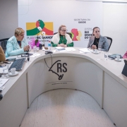 A foto mostra um grupo de oito pessoas, entre homens e mulheres, sentadas em torno de uma mesa branca em formato de U, participando de uma reunião. No centro da mesa, com uma blusa verde e óculos, está a Secretária da Saúde, Arita Bergmann, atenta à discussão. Os participantes estão vestidos com trajes formais e casuais, com papéis, notebooks e xícaras de café na mesa. Ao fundo, uma parede clara com banners da "Secretaria da Saúde" e "Governo do Estado" indica o contexto institucional do encontro.