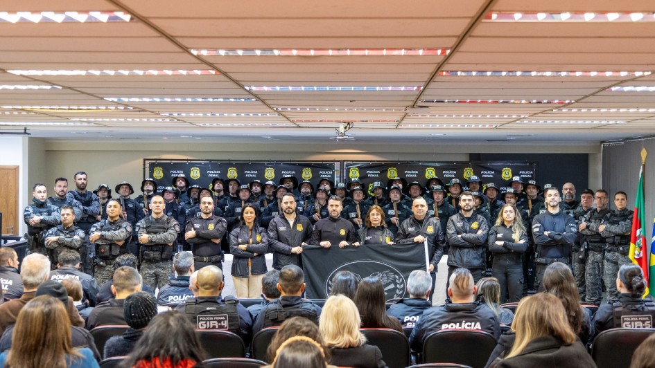 Um grupo numeroso de agentes da Polícia Penal posa uniformizado para foto em um auditório. Na frente, uma fileira de homens e mulheres segurando uma bandeira preta com o brasão da Polícia Penal. Ao fundo, mais agentes alinhados com escudos e capacetes. Sentados de costas, diversos outros policiais acompanham a cerimônia. À direita, estão posicionadas as bandeiras do Brasil, do Rio Grande do Sul e institucional.