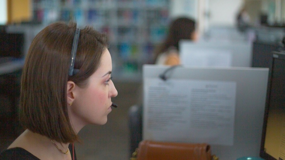 A foto mostra uma mulher jovem com cabelo castanho-claro, usando um headset preto e um microfone perto da boca, sentada em uma mesa de escritório e olhando para a direita. Ela tem um brinco dourado e um colar fino no pescoço. No fundo, há um ambiente de escritório com baias e, desfocado, um armário com prateleiras cheias de livros ou pastas coloridas. Outras pessoas, também desfocadas, são visíveis ao fundo, trabalhando em computadores. A iluminação é clara e difusa, vinda do teto.