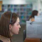 A foto mostra uma mulher jovem com cabelo castanho-claro, usando um headset preto e um microfone perto da boca, sentada em uma mesa de escritório e olhando para a direita. Ela tem um brinco dourado e um colar fino no pescoço. No fundo, há um ambiente de escritório com baias e, desfocado, um armário com prateleiras cheias de livros ou pastas coloridas. Outras pessoas, também desfocadas, são visíveis ao fundo, trabalhando em computadores. A iluminação é clara e difusa, vinda do teto.