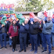 Imagem ao ar livre. Diversas pessoas estão sorrindo para a foto, em pé, segurando sacas de sementes nas cores verde, vermelha e azul. 
