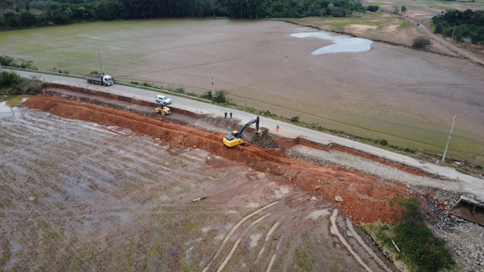 Imagem aérea mostra uma obra de reconstrução em andamento em uma estrada rural, que parece ter sido danificada por enxurradas ou alagamentos. À esquerda da imagem, observa-se um campo alagado e com marcas de pneus na terra encharcada. Ao centro, há uma escavadeira amarela trabalhando em uma grande vala aberta ao lado da pista, com presença de caminhão basculante, trator e trabalhadores com coletes de segurança. A estrada está parcialmente interditada com cones, e há alguns veículos estacionados próximos ao trecho em obras. Ao fundo, há áreas agrícolas e vegetação.