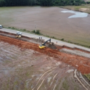 Imagem aérea mostra uma obra de reconstrução em andamento em uma estrada rural, que parece ter sido danificada por enxurradas ou alagamentos. À esquerda da imagem, observa-se um campo alagado e com marcas de pneus na terra encharcada. Ao centro, há uma escavadeira amarela trabalhando em uma grande vala aberta ao lado da pista, com presença de caminhão basculante, trator e trabalhadores com coletes de segurança. A estrada está parcialmente interditada com cones, e há alguns veículos estacionados próximos ao trecho em obras. Ao fundo, há áreas agrícolas e vegetação.