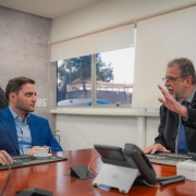 Em uma sala de reuniões, dois homens estão sentados à mesa. Ganbriel, de terno azul, ouve atentamente. O CEO da Taurus, de terno escuro e gravata azul, gesticula com a mão levantada enquanto fala. Há telefones e papéis sobre a mesa.