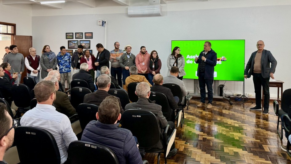 A foto mostra o Secretário da Agricultura, Edivilson Brum, um homem de pele clara e cabelo escuro, em pé no centro da imagem. Ele veste uma jaqueta escura e está segurando um microfone, falando para uma plateia. Ao seu lado direito, um telão verde exibe alguns logotipos. À esquerda do secretário, há um grupo de cerca de dez pessoas em pé, incluindo homens e mulheres, olhando para a plateia. Em primeiro plano, uma plateia de aproximadamente trinta pessoas, a maioria homens de meia-idade, está sentada em cadeiras escuras, com muitos de costas para a câmera. O ambiente é uma sala ampla com teto branco e iluminação fluorescente, e no fundo, uma porta de madeira e quadros na parede.