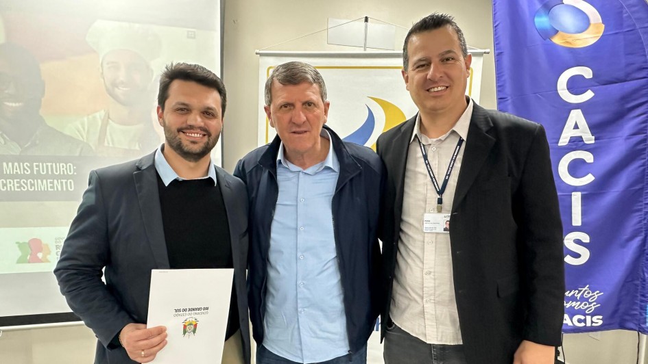 Foto posada de frente de três homens em trajes formais de pé, num ambiente fechado de paredes claras. Eles sorriem, e o da esquerda segura um documento.