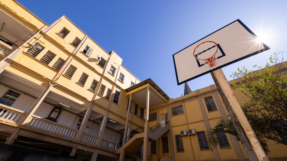 Imagem externa. Há uma cesta de basquete em destaque no lado direito da foto. No lado esquerdo, aparece a estrutura do colégio, com três pisos, em tons amarelos. 