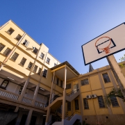 Imagem externa. Há uma cesta de basquete em destaque no lado direito da foto. No lado esquerdo, aparece a estrutura do colégio, com três pisos, em tons amarelos. 