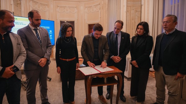A foto mostra um grupo de seis pessoas em pé ao redor de uma pequena mesa de madeira, em um ambiente interno formal. No centro, o governador em exercício, Gabriel Souza, com barba, vestido com um terno marrom e camisa escura, está assinando um documento. Ele está inclinado sobre a mesa, com a caneta na mão, e todos os demais o observam com atenção. Ao lado esquerdo de Gabriel, uma mulher de cabelo escuro e camisa preta está ao seu lado. O resto do grupo é composto por quatro homens de terno. O ambiente tem paredes claras e uma janela alta com cortinas. Ao fundo, um telão mostra uma apresentação e, à direita, há uma estátua e uma coluna. O chão é de madeira.