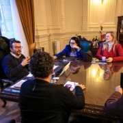 A foto mostra quatro pessoas sentadas em uma mesa de reunião grande e retangular, com tampo de madeira, em uma sala com arquitetura clássica. A secretária do Meio Ambiente e Infraestrutura, Marjorie Kauffmann, está sentada à frente e à esquerda, vestindo um casaco azul e com o cotovelo apoiado na mesa, olhando para o lado. À sua frente, um homem de blusa escura e óculos, que é o Chefe da Casa Civil, Artur Lemos, está sentado na ponta da mesa. Do outro lado da mesa, um homem com barba e óculos, com um suéter azul, está sentado à esquerda e interagindo. Uma quarta mulher de blusa vermelha e cabelos loiros está sentada à direita. A mesa tem papéis, tablets, xícaras de café e notebooks. A sala tem teto com adornos, janelas com cortinas longas e paredes com molduras. 