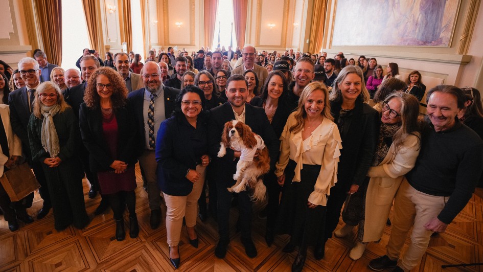 A foto mostra um grupo grande e diverso de pessoas, incluindo homens e mulheres, posando para uma foto em um salão espaçoso com um teto alto e um grande afresco em uma das paredes. No centro, o governador em exercício, Gabriel Souza, está segurando um cão de porte médio, de pelagem branca e caramelo, que veste uma camiseta branca. Ao lado de Gabriel, há um grupo de pessoas sorridentes, incluindo a Secretária do Meio Ambiente e Infraestrutura, Marjorie Kauffmann, de blusa branca. A plateia, formada por muitas pessoas, está em pé no fundo. O piso é de madeira e a iluminação é natural.