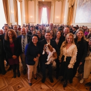 A foto mostra um grupo grande e diverso de pessoas, incluindo homens e mulheres, posando para uma foto em um salão espaçoso com um teto alto e um grande afresco em uma das paredes. No centro, o governador em exercício, Gabriel Souza, está segurando um cão de porte médio, de pelagem branca e caramelo, que veste uma camiseta branca. Ao lado de Gabriel, há um grupo de pessoas sorridentes, incluindo a Secretária do Meio Ambiente e Infraestrutura, Marjorie Kauffmann, de blusa branca. A plateia, formada por muitas pessoas, está em pé no fundo. O piso é de madeira e a iluminação é natural.