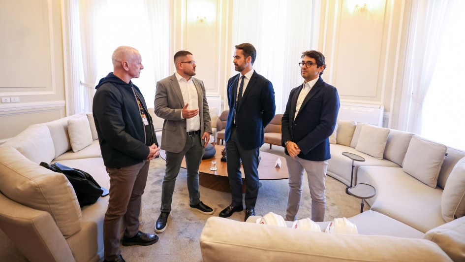 A foto mostra quatro homens em pé em um salão com paredes claras e janelas grandes. No centro, o governador Eduardo Leite veste um terno azul-marinho com uma gravata azul-clara. À esquerda dele, de terno cinza, está o vice-presidente do Conselho de Gestão do Internacional, Victor Grunberg, que gesticula enquanto fala. Ao lado do governador, o sócio-fundador da Tornak Holding, Fernando Tornaim, veste um terno escuro. À esquerda, o secretário de Comunicação, Caio Tomazeli, está de casaco preto e calça marrom. Eles estão em pé, próximos a sofás brancos.