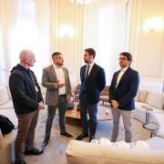 A foto mostra quatro homens em pé em um salão com paredes claras e janelas grandes. No centro, o governador Eduardo Leite veste um terno azul-marinho com uma gravata azul-clara. À esquerda dele, de terno cinza, está o vice-presidente do Conselho de Gestão do Internacional, Victor Grunberg, que gesticula enquanto fala. Ao lado do governador, o sócio-fundador da Tornak Holding, Fernando Tornaim, veste um terno escuro. À esquerda, o secretário de Comunicação, Caio Tomazeli, está de casaco preto e calça marrom. Eles estão em pé, próximos a sofás brancos.