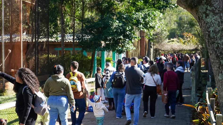 A imagem mostra um grupo grande de pessoas caminhando por um caminho pavimentado dentro de um zoológico ou parque natural. À esquerda, há uma cerca de tela com um recinto de animais, onde algumas pessoas — incluindo uma mulher com cabelo cacheado e blusa preta — estão apontando, possivelmente mostrando um animal para as crianças.

O local é arborizado, com sombra de árvores projetada no chão. Entre os visitantes, há adultos e crianças, muitas com mochilas e roupas casuais. Um menino pequeno, de boné colorido, segura a mão de um adulto enquanto caminha. Ao fundo, o caminho continua e está bastante movimentado, indicando grande fluxo de visitantes no local. O clima parece agradável e ensolarado.