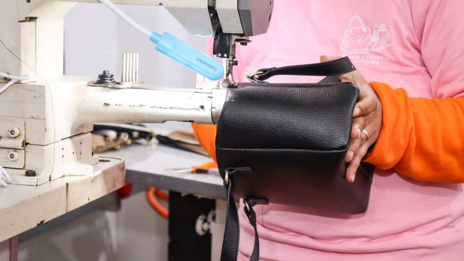 A foto mostra a parte superior do corpo de uma apenada, vestindo uma camiseta rosa por cima de uma blusa de manga comprida laranja, sentada em frente a uma máquina de costura industrial. Ela está costurando uma pequena bolsa preta de couro sintético, com alças pretas. A máquina de costura tem um braço articulado com uma luz azul na ponta, iluminando a área de trabalho. A mulher usa um anel no dedo anular da mão direita. A foto foca na atividade de costura, com o restante do ambiente desfocado, indicando uma oficina ou linha de produção.