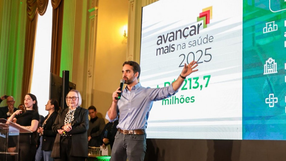 Em um auditório iluminado, o governador do Rio Grande do Sul fala ao microfone diante de um grande telão que exibe o texto “Avançar Mais na Saúde 2025 – R$ 213,7 milhões”. Ele está de camisa azul e calça jeans, com um dos braços levantado em gesto de ênfase. Ao fundo, outras pessoas acompanham, incluindo uma mulher de óculos e cabelo grisalho curto, que também segura um microfone. À esquerda, uma intérprete de Libras traduz o discurso.