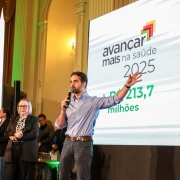 Em um auditório iluminado, o governador do Rio Grande do Sul fala ao microfone diante de um grande telão que exibe o texto “Avançar Mais na Saúde 2025 – R$ 213,7 milhões”. Ele está de camisa azul e calça jeans, com um dos braços levantado em gesto de ênfase. Ao fundo, outras pessoas acompanham, incluindo uma mulher de óculos e cabelo grisalho curto, que também segura um microfone. À esquerda, uma intérprete de Libras traduz o discurso.