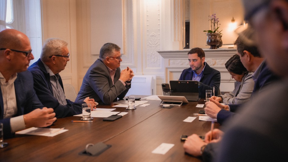 Foto em sala de reunião, com seis pessoas ao redor de uma mesa retangular de madeira. Gabriel aparece ao centro da imagem, conversando com os presentes.