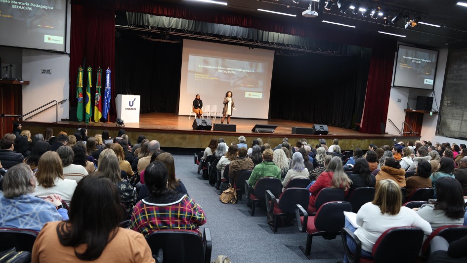 Fotografia de um auditório lotado, com dezenas de pessoas sentadas, voltadas para o palco. No centro do palco, uma mulher está em pé, falando ao público, enquanto outra pessoa está sentada mais ao fundo. Há duas grandes telas projetando a mesma apresentação, com o título “Formação Mentoria Pedagógica SEDUC/RS”. À esquerda do palco, estão posicionadas bandeiras do Brasil, do Rio Grande do Sul e de outras instituições, além do logotipo da Unisinos. As luzes do teto iluminam a cena, criando um ambiente formal de capacitação e palestra.