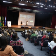 Fotografia de um auditório lotado, com dezenas de pessoas sentadas, voltadas para o palco. No centro do palco, uma mulher está em pé, falando ao público, enquanto outra pessoa está sentada mais ao fundo. Há duas grandes telas projetando a mesma apresentação, com o título “Formação Mentoria Pedagógica SEDUC/RS”. À esquerda do palco, estão posicionadas bandeiras do Brasil, do Rio Grande do Sul e de outras instituições, além do logotipo da Unisinos. As luzes do teto iluminam a cena, criando um ambiente formal de capacitação e palestra.