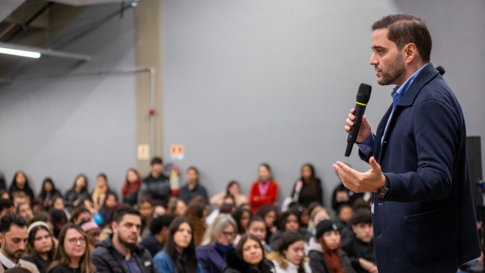 Uma pessoa está de pé, segurando um microfone e falando para um público grande e atento numa sala com iluminação industrial. Há um extintor de incêndio na parede ao fundo.