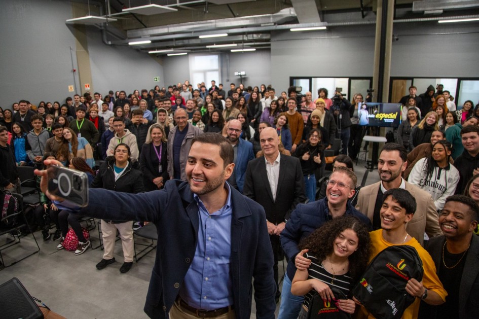 Um grupo grande de pessoas reunido num espaço interno, estilo conferência. Um homem está tirando uma selfie com a multidão ao fundo. Algumas pessoas usam crachás e há uma tela com a palavra “especial” visível.