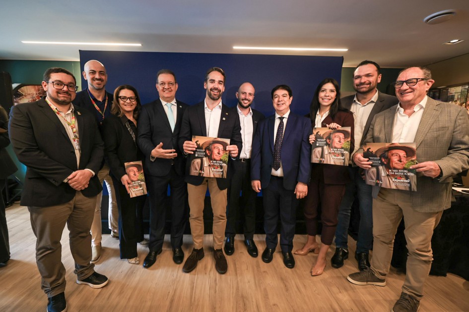 Esta é uma fotografia de um evento formal em um salão moderno, onde um homem está em pé discursando para uma plateia.
No primeiro plano à direita, um homem branco com barba, vestindo um blazer azul-marinho, camisa social clara e calças cáqui, segura um microfone branco com a mão direita e gesticula com a esquerda. Ele olha para a plateia.
À esquerda do orador, em primeiro plano, vários participantes estão sentados em poltronas escuras. Ao centro, um homem de óculos e terno escuro está sentado, com uma expressão atenta.
Atrás da primeira fila, uma grande plateia está em pé, formando uma multidão que se estende por todo o fundo da sala. Muitas pessoas estão com celulares e câmeras, registrando o evento.
O ambiente é amplo, com paredes brancas e janelas grandes ao fundo. Na parede à direita do orador, um painel preto exibe um logotipo com silhuetas coloridas de cabeças humanas. A iluminação é clara e a foto é nítida.