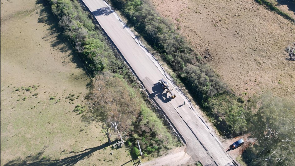 Imagem aérea mostra uma estrada em obras localizada em uma área rural. A via, cercada por vegetação rasteira e árvores de ambos os lados, está sendo pavimentada. No centro da imagem, há uma máquina pesada (possivelmente uma motoniveladora ou pavimentadora) em operação, com um trabalhador próximo a ela. À direita, fora da estrada, vê-se um carro estacionado em uma área de terra batida. A paisagem ao redor é composta por campos abertos e áreas de mata esparsa. A imagem foi tirada sob luz do dia, com sombras projetadas pelas árvores indicando sol forte.